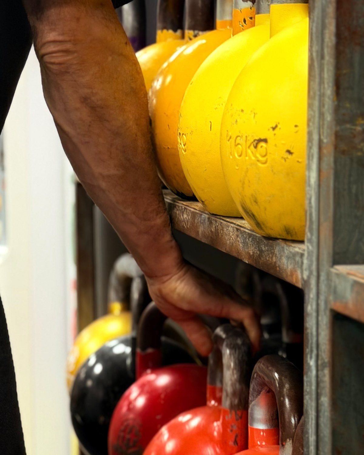Mano cogiendo una kettlebell del estante del centro, con kettlebells amarillas, rojas y naranjas